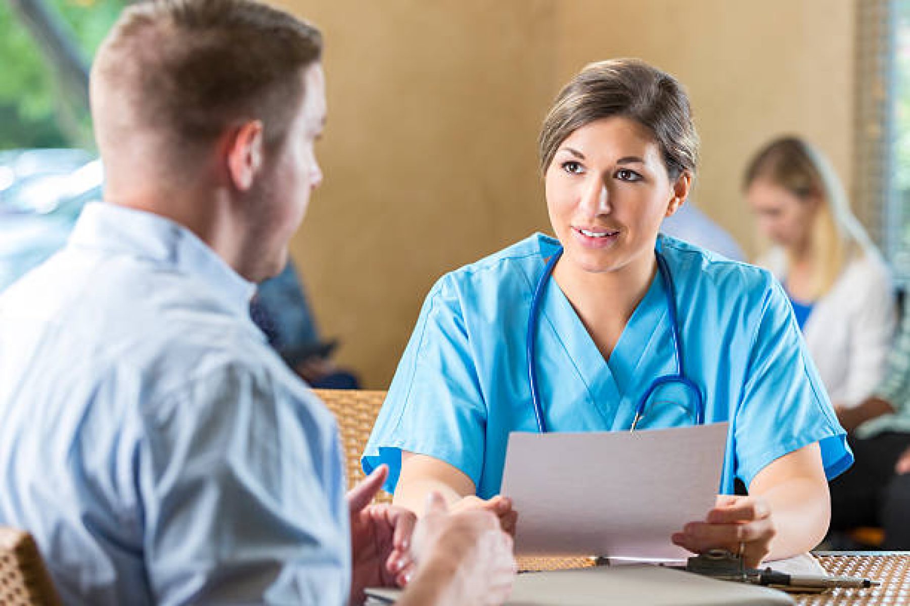 Mid adult Caucasian woman is manager of nursing staff at hospital. She is reading a resume and interviewing a male candidate for a healthcare job. Other potential employees are waiting to be interviewed during hospital's staff recruitment event.